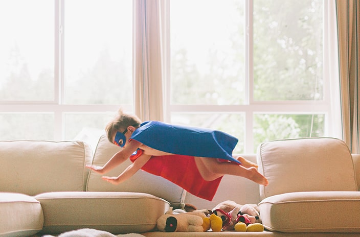 Boy with superhero cape flying over stuffed animals on couch.