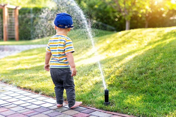 Toddler standing next to lawn sprinkler.